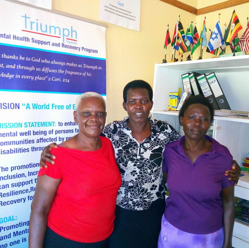 three ugandan woman stand next to a sign for Triumph Uganda