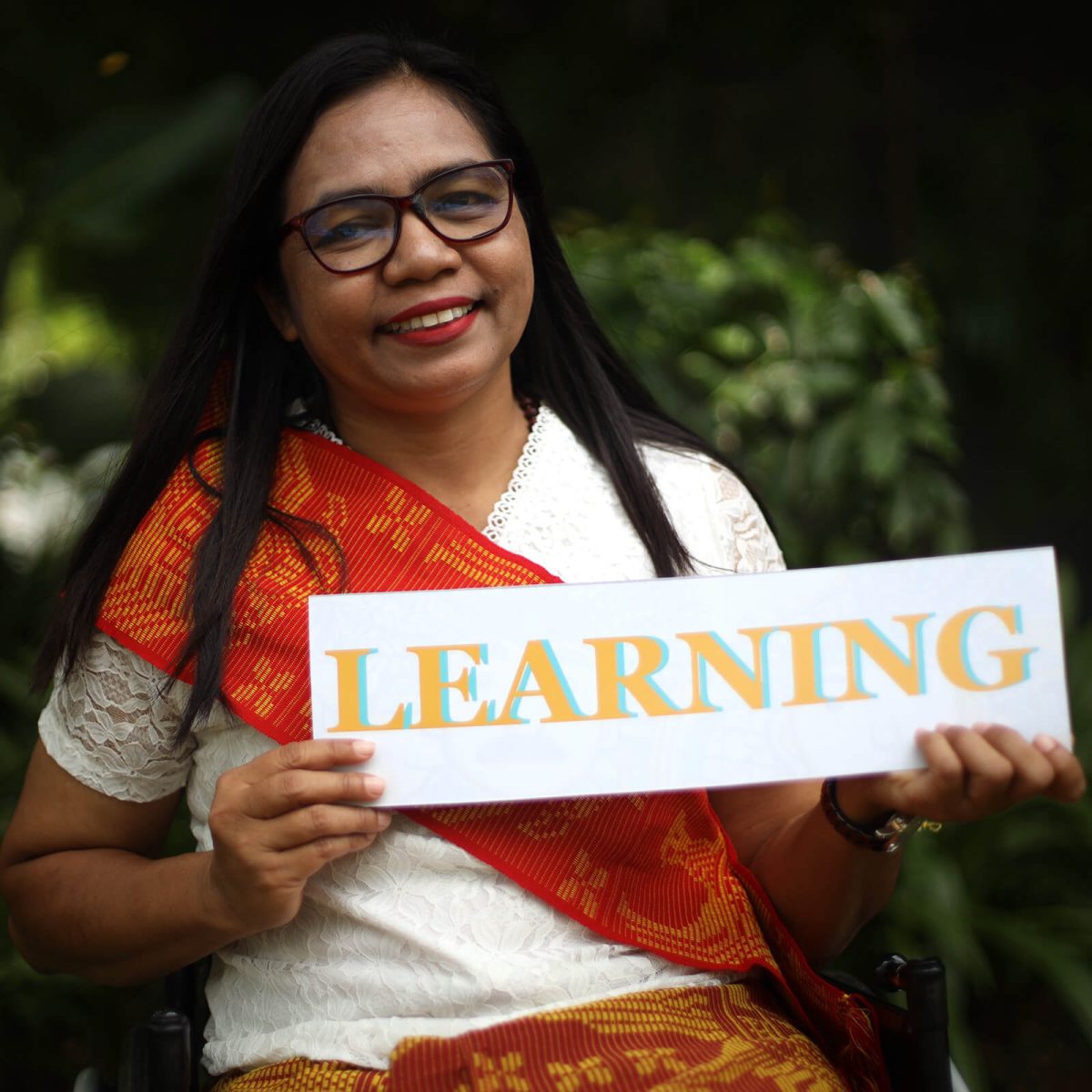 Photo of an Indonesian woman smiling and wearing dark glasses. She has long dark hair and is wearing a traditional dress while holding a sign: LEARNING.