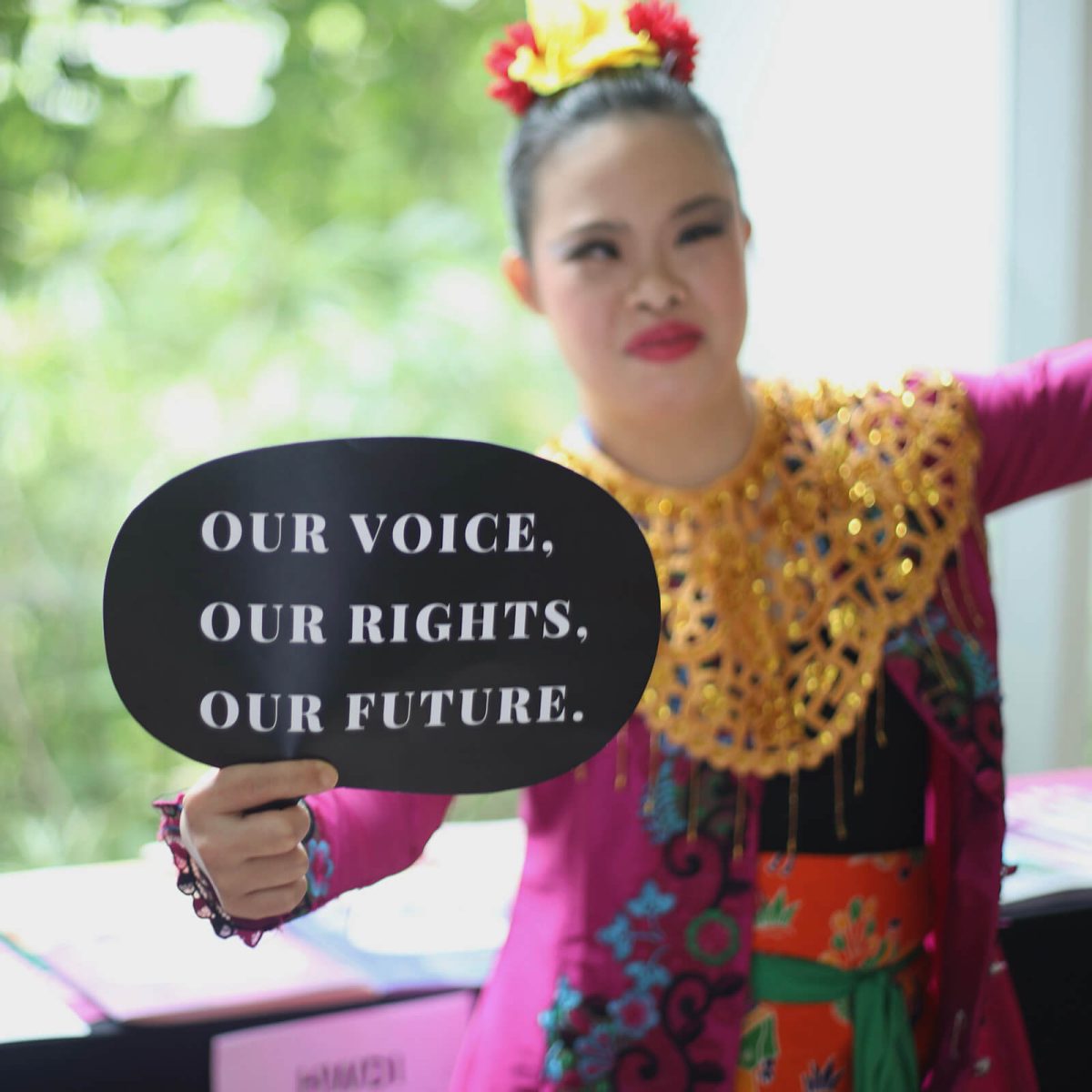 A young Indonesian disability rights activist holds a sign: Our Voice. Our Rights. Our Future. She’s wearing a bright pink traditional tunic with a gold necklace and flowers on her hair.