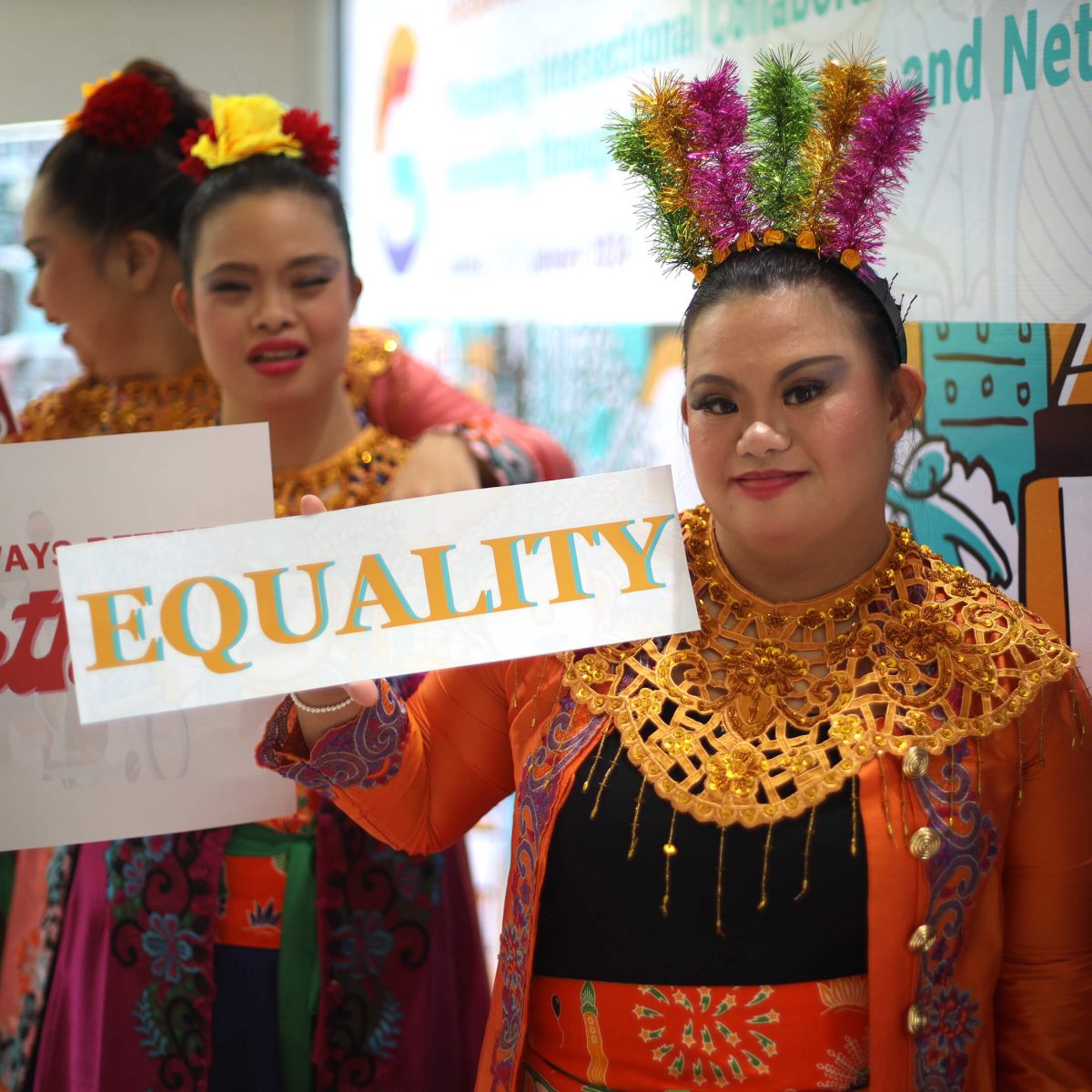 Three women are wearing bright traditional Indonesian dresses. A young woman in front is holding a sign, 'Equality.'