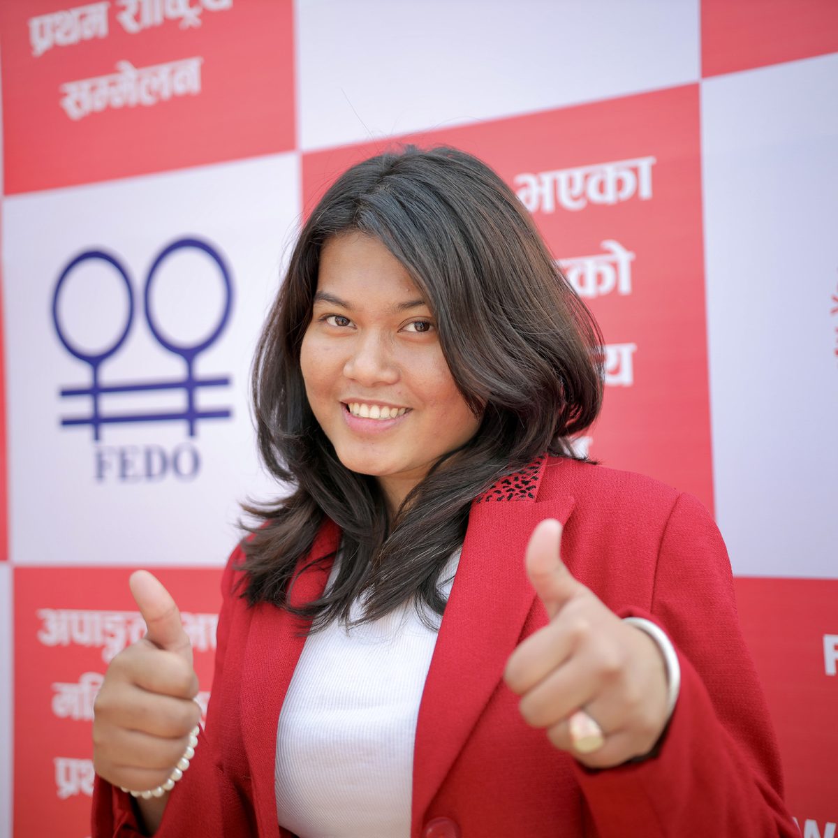 A young Nepali woman with short dark hair is smiling and giving a thumbs up. She's wearing a red jacket and is standing in front of a banner.