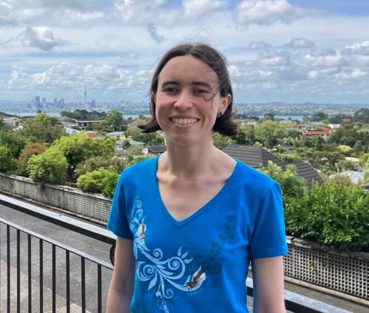 Áine stands smiling outdoors on a house balcony, their short brown hair blowing in the summer breeze. Suburban treetops dominate the background, with Auckland's skyline visible in the distance.