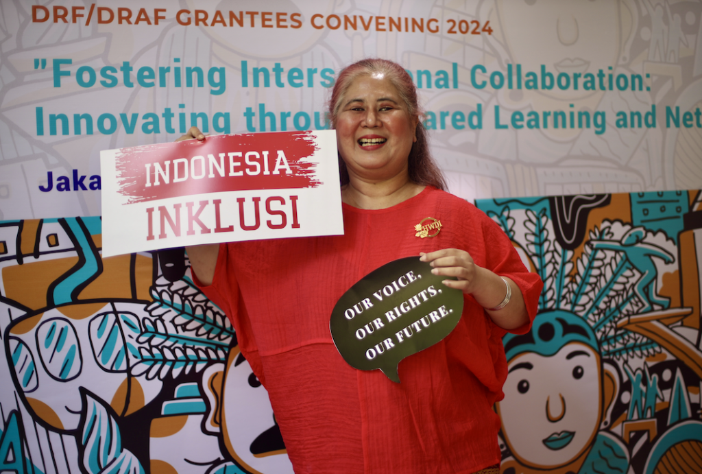 A woman wearing a red top is smiling and holding signs: Indonesia INKLUSI and Our Voices, Our Rights.