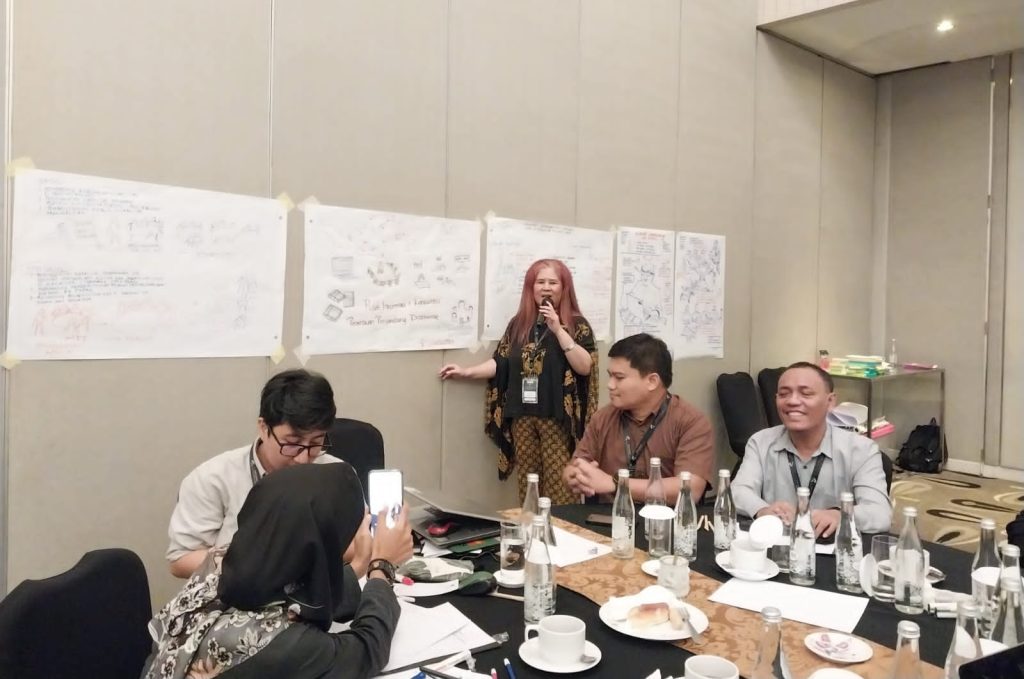 A woman is standing and speaking into a microphone in a room with people seated around tables and there are flip charts on the wall. 