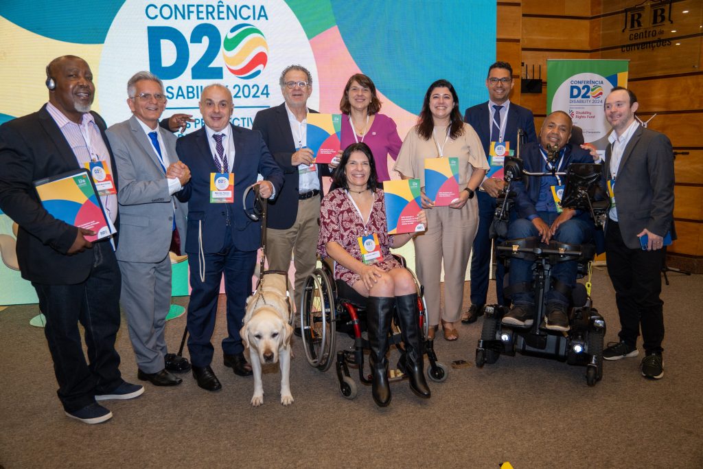 A group of people posing together at the D20 Conference inside a room. They are standing in front of the banner with text: "Conferência D20 - Sabia 2024." The group includes men and women with diverse disabilities.