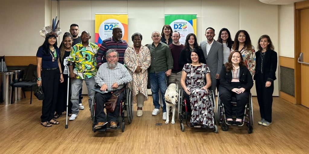 Two colorful banners with the D20 logo and conference details are displayed behind a group of people. The group includes individuals with disabilities, some are using wheelchairs, a guide dog, and a person using a crutch. 