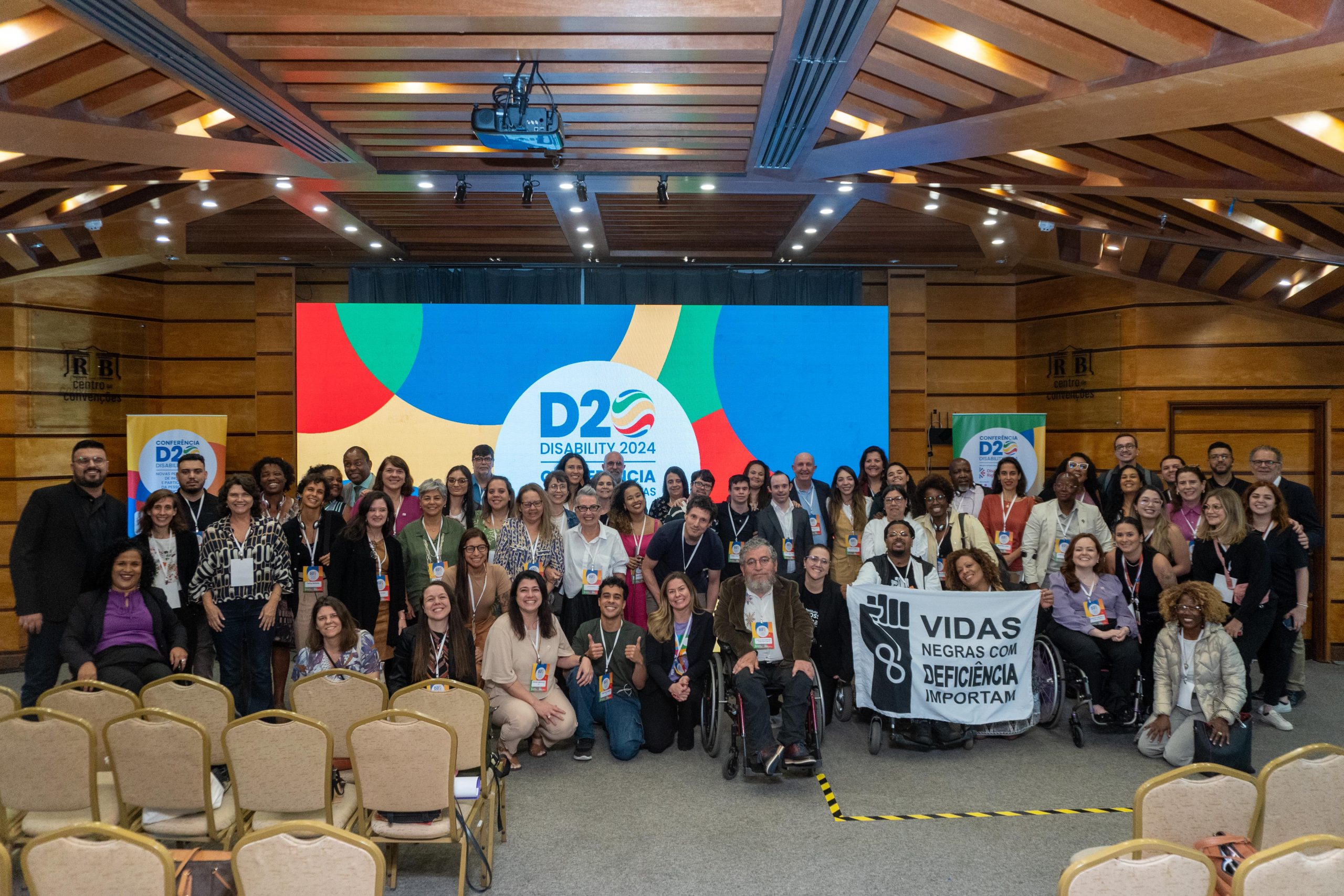 A group of people are gathered inside a room. Behind them is a banana with text: D 20 Disability 2024.