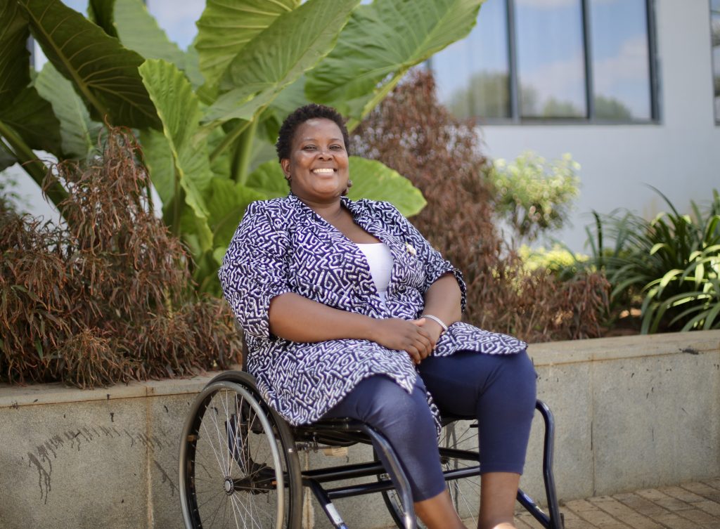 An African woman is smiling outdoors. She's using a wheelchair and is wearing a blue and white printed top and blue pants.