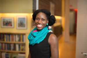 A headshot of Antoinette, a Black woman wearing a blue scarf and a black top, smiling indoors.