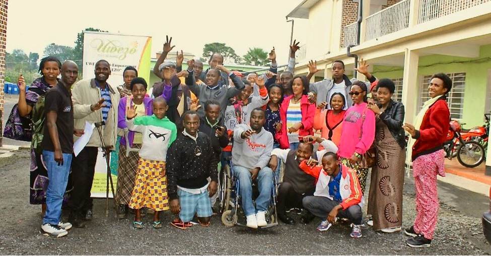 A group of young people with disabilities smile and many of them wave.