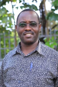 Francis Xavier, wearing a grey printed shirt and glasses, stands outside.