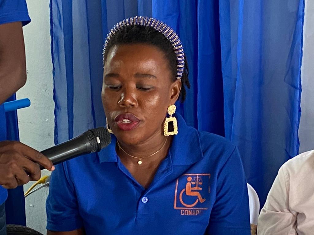 A picture of a woman wearing a blue t-shirt with a wheelchair design speaking in a microphone