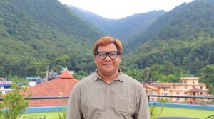 Krishna, wearing glasses, smiles. Behind him is a view of a mountain range
