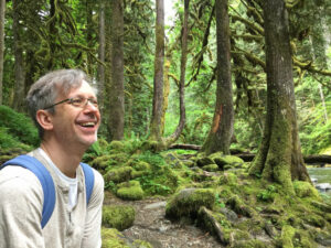 An outdoor shot of Michael Szporluck in a lush green forest in Oregon. He is smiling and wearing glasses.