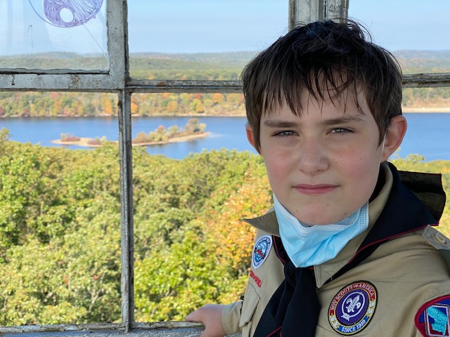 Sebastian, a 12 year old boy, looks at the camera smiling with his face mask pulled down. He is standing in front of a window that looks out over water and trees.