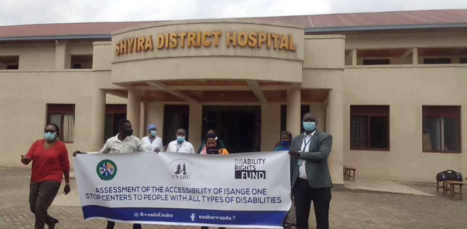 7 people walk holding a banner reading Assessment of the accessibility of isange one stop centers to people with all types of disabilities'. The district hospital is behind them.