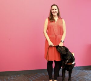 Photo of Kerry standing in front of a pink wall with her guide dog, Miles, a Black Labrador.
