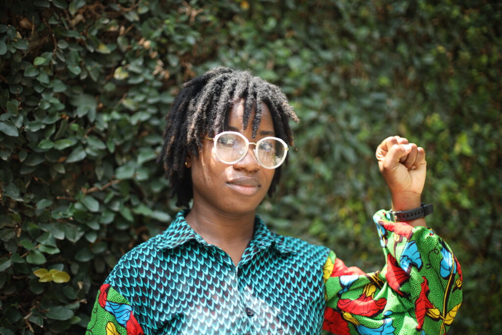 Benedicta stands up with her fist raised with green foliage in the background.