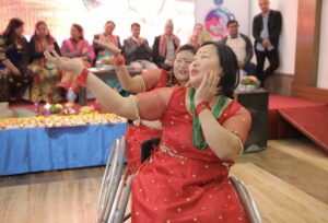 A photo of two Nepali indigenous women in red dresses doing a traditional dance. They are using wheelchairs.